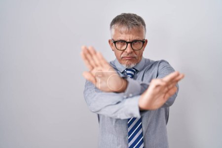 Foto de Hombre de negocios hispano con cabello gris usando gafas expresión de rechazo cruzando brazos y palmas haciendo signo negativo, cara enojada - Imagen libre de derechos