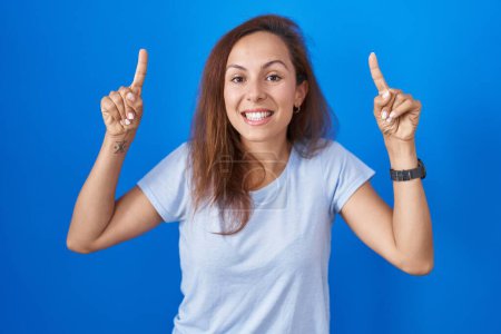 Photo for Brunette woman standing over blue background smiling amazed and surprised and pointing up with fingers and raised arms. - Royalty Free Image