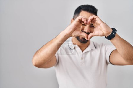 Téléchargez les photos : Jeune homme hispanique avec barbe portant des vêtements décontractés sur fond blanc faisant forme de coeur avec la main et les doigts souriant en regardant à travers signe - en image libre de droit