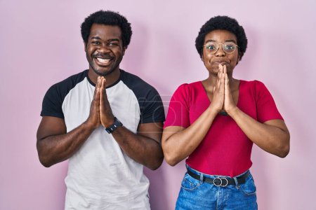 Photo for Young african american couple standing over pink background praying with hands together asking for forgiveness smiling confident. - Royalty Free Image