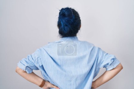 Téléchargez les photos : Jeune fille moderne avec les cheveux bleus debout sur fond blanc debout vers l'arrière regardant loin avec les bras sur le corps - en image libre de droit