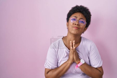 Foto de Joven mujer afroamericana de pie sobre fondo rosa mendigando y orando con las manos juntas con la esperanza expresión en la cara muy emocional y preocupado. mendicidad. - Imagen libre de derechos