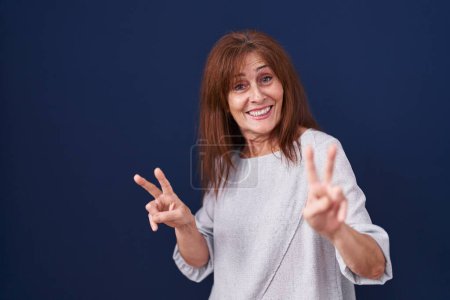 Téléchargez les photos : Femme d'âge moyen debout sur fond bleu souriant en regardant la caméra montrant les doigts faisant signe de victoire. numéro deux. - en image libre de droit