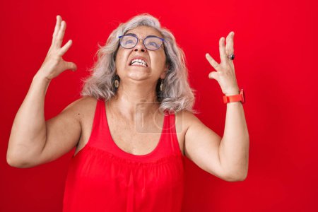 Photo for Middle age woman with grey hair standing over red background crazy and mad shouting and yelling with aggressive expression and arms raised. frustration concept. - Royalty Free Image