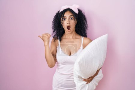 Photo for Hispanic woman with curly hair wearing sleep mask and pajama holding pillow surprised pointing with hand finger to the side, open mouth amazed expression. - Royalty Free Image