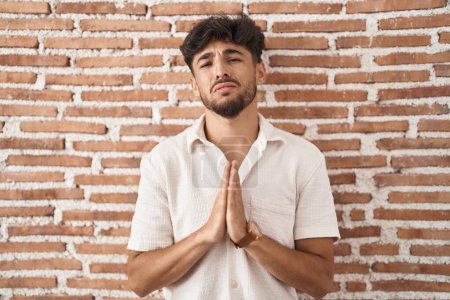 Photo for Arab man with beard standing over bricks wall background begging and praying with hands together with hope expression on face very emotional and worried. begging. - Royalty Free Image