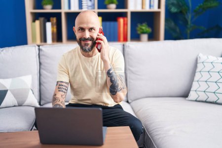 Téléchargez les photos : Young bald man talking on smartphone using laptop at home - en image libre de droit