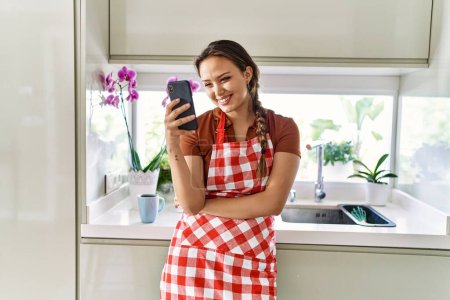 Téléchargez les photos : Jeune belle femme hispanique portant tablier en utilisant un smartphone à la cuisine - en image libre de droit