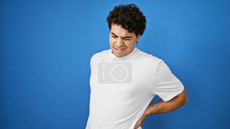 Téléchargez les photos : Jeune homme latin souffrant de maux de dos debout sur fond bleu isolé - en image libre de droit