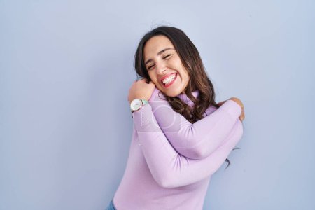 Téléchargez les photos : Jeune femme brune debout sur fond bleu se câlinant heureux et positif, souriant confiant. amour de soi et soins de soi - en image libre de droit
