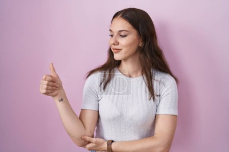 Photo for Young hispanic girl standing over pink background looking proud, smiling doing thumbs up gesture to the side - Royalty Free Image