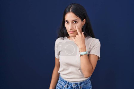Téléchargez les photos : Jeune femme hispanique debout sur fond bleu pointant vers l'œil vous regardant geste, expression suspecte - en image libre de droit