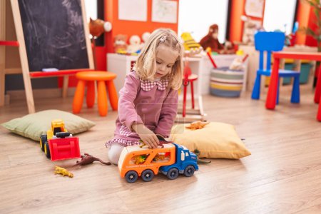 Foto de Adorable chica rubia jugando con el juguete del camión y dinosaurio sentado en el suelo en el jardín de infantes - Imagen libre de derechos