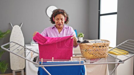 Photo for Young beautiful latin woman listening to music hanging clothes on clothesline at laundry room - Royalty Free Image