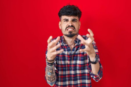 Photo for Young hispanic man with beard standing over red background shouting frustrated with rage, hands trying to strangle, yelling mad - Royalty Free Image