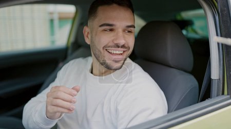 Téléchargez les photos : Jeune homme hispanique souriant confiant assis sur la voiture à la rue - en image libre de droit