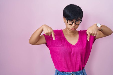 Photo for Young asian woman with short hair standing over pink background pointing down looking sad and upset, indicating direction with fingers, unhappy and depressed. - Royalty Free Image
