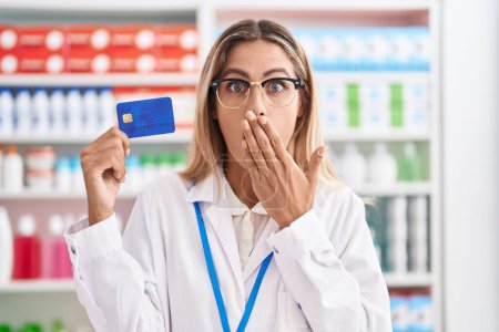 Foto de Mujer rubia joven que trabaja en la farmacia sosteniendo la tarjeta de crédito que cubre la boca con la mano, conmocionada y asustada por el error. expresión sorprendida - Imagen libre de derechos