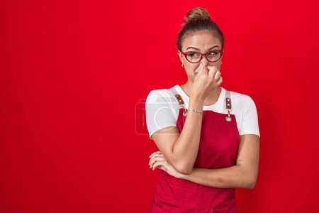 Téléchargez les photos : Jeune femme hispanique portant tablier de serveuse sur fond rouge sentant quelque chose de puant et dégoûtant, odeur intolérable, retenant souffle avec les doigts sur le nez. mauvaise odeur - en image libre de droit