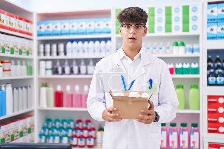 Téléchargez les photos : Adolescent hispanique travaillant à la pharmacie drugstore tenant boîte avec des pilules peur et étonné avec la bouche ouverte pour la surprise, visage incrédule - en image libre de droit