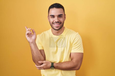 Foto de Joven hispano de pie sobre fondo amarillo con una gran sonrisa en la cara, señalando con el dedo de la mano hacia un lado mirando a la cámara. - Imagen libre de derechos