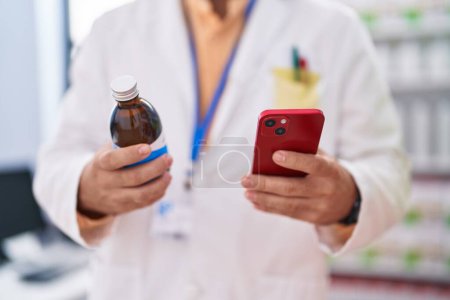 Téléchargez les photos : Young hispanic man pharmacist using smartphone holding medication bottle at pharmacy - en image libre de droit