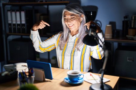 Foto de Mujer de mediana edad con el pelo gris trabajando en la oficina por la noche mirando confiado con sonrisa en la cara, señalándose con los dedos orgullosos y felices. - Imagen libre de derechos
