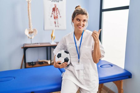 Téléchargez les photos : Jeune femme travaillant à la clinique de thérapie de football pointant le pouce vers le côté souriant heureux avec la bouche ouverte - en image libre de droit