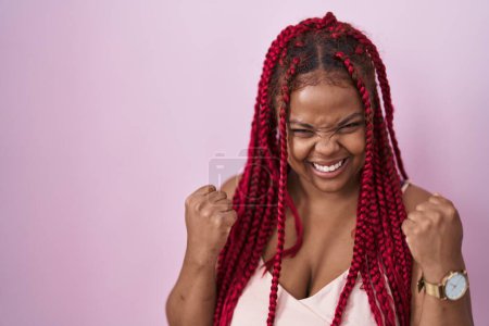 Téléchargez les photos : Femme afro-américaine avec les cheveux tressés debout sur fond rose très heureux et excité de faire geste gagnant avec les bras levés, souriant et criant pour le succès. concept de célébration. - en image libre de droit
