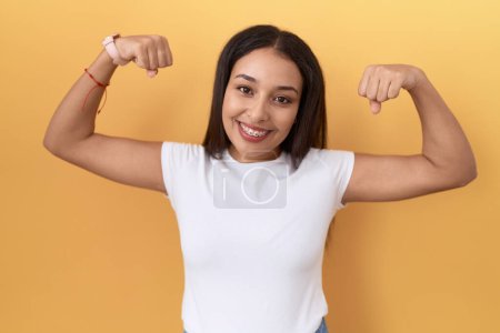Téléchargez les photos : Jeune femme arabe portant un t-shirt blanc décontracté sur fond jaune montrant les muscles des bras souriant fier. concept de fitness. - en image libre de droit