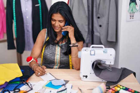 Téléchargez les photos : Moyen Age femme hispanique tailleur parler sur smartphone écriture sur ordinateur portable au magasin de tailleur - en image libre de droit