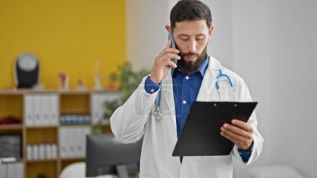 Photo for Young hispanic man doctor reading document on clipboard talking on smartphone at the clinic - Royalty Free Image