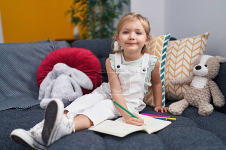 Photo for Adorable blonde girl preschool student sitting on sofa drawing on notebook at home - Royalty Free Image
