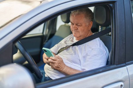 Téléchargez les photos : Moyen âge homme aux cheveux gris en utilisant smartphone assis sur la voiture à la rue - en image libre de droit