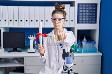 Photo for Teenager girl working at scientist laboratory holding degree pointing with finger to the camera and to you, confident gesture looking serious - Royalty Free Image