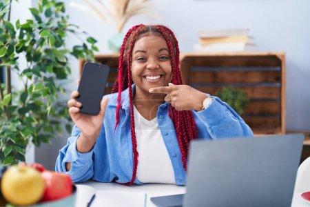 Téléchargez les photos : Femme afro-américaine avec des cheveux tressés tenant smartphone montrant écran vierge souriant heureux pointant avec la main et le doigt - en image libre de droit