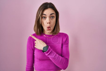 Foto de Mujer hispana de pie sobre fondo rosa sorprendida señalando con el dedo hacia un lado, boca abierta expresión asombrada. - Imagen libre de derechos