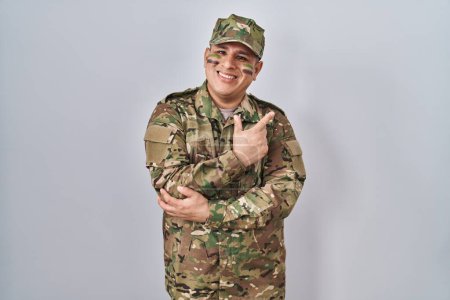 Téléchargez les photos : Jeune homme hispanique portant l'uniforme de l'armée de camouflage avec un grand sourire sur le visage, pointant avec la main et le doigt vers le côté regardant la caméra. - en image libre de droit