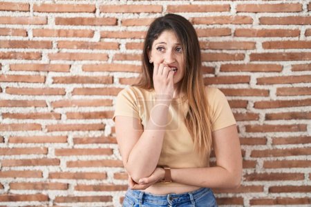 Photo for Young brunette woman standing over bricks wall looking stressed and nervous with hands on mouth biting nails. anxiety problem. - Royalty Free Image
