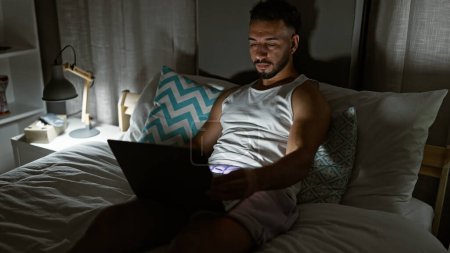Téléchargez les photos : Jeune homme arabe utilisant un ordinateur portable assis sur le lit dans la chambre - en image libre de droit
