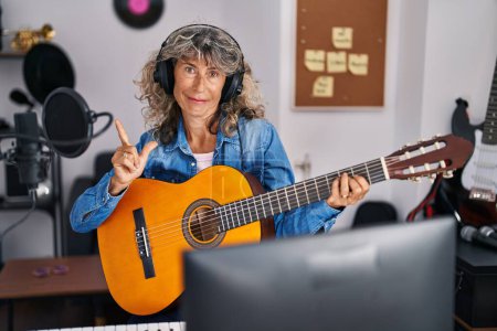 Foto de Mujer de mediana edad tocando la guitarra clásica en el estudio de música sonriendo feliz señalando con la mano y el dedo a un lado - Imagen libre de derechos