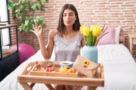 Photo for Brunette young woman eating breakfast sitting on the bed relax and smiling with eyes closed doing meditation gesture with fingers. yoga concept. - Royalty Free Image