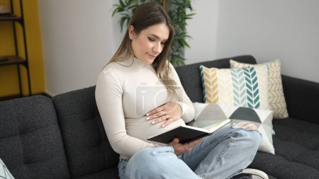 Foto de Joven embarazada leyendo libro tocando vientre en casa - Imagen libre de derechos