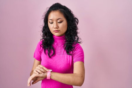 Foto de Joven mujer asiática de pie sobre fondo rosa comprobar el tiempo en reloj de pulsera, relajado y seguro - Imagen libre de derechos