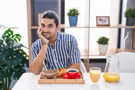 Téléchargez les photos : Homme hispanique avec les cheveux longs assis sur la table ayant le petit déjeuner pensant fatigué et ennuyé avec des problèmes de dépression avec les bras croisés. - en image libre de droit