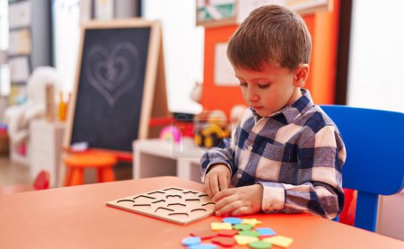 Foto de Adorable chico caucásico jugando con matemáticas juego de puzzle sentado en la mesa en el jardín de infantes - Imagen libre de derechos