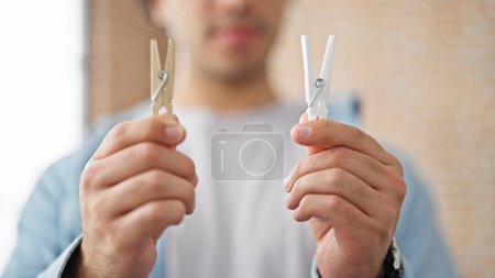 Photo for Young latin man choosing wooden or plastic clothespin thinking at home - Royalty Free Image