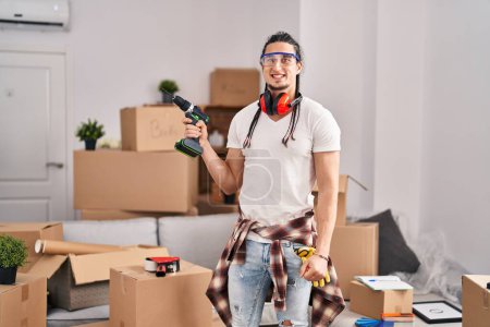 Photo for Hispanic man with long hair holding screwdriver at new home looking positive and happy standing and smiling with a confident smile showing teeth - Royalty Free Image