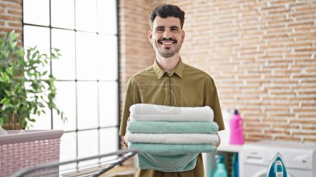 Téléchargez les photos : Jeune homme hispanique souriant confiant tenant serviettes pliées à la buanderie - en image libre de droit
