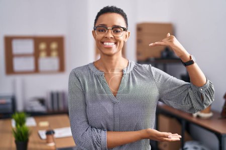 Téléchargez les photos : Femme afro-américaine travaillant au bureau portant des lunettes geste avec les mains montrant grand et grand signe de taille, symbole de mesure. souriant en regardant la caméra. concept de mesure. - en image libre de droit